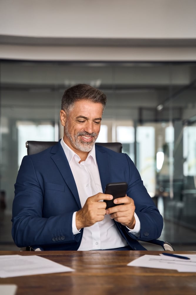 Geschäftsmann und CEO sitzt lächelnd in einem Büro und arbeitet gleichzeitig mit Laptop und Smartphone. Der CEO oder Manager hält ein Mobiltelefon in der Hand, nutzt eine App und arbeitet online an seinem modernen Arbeitsplatz.